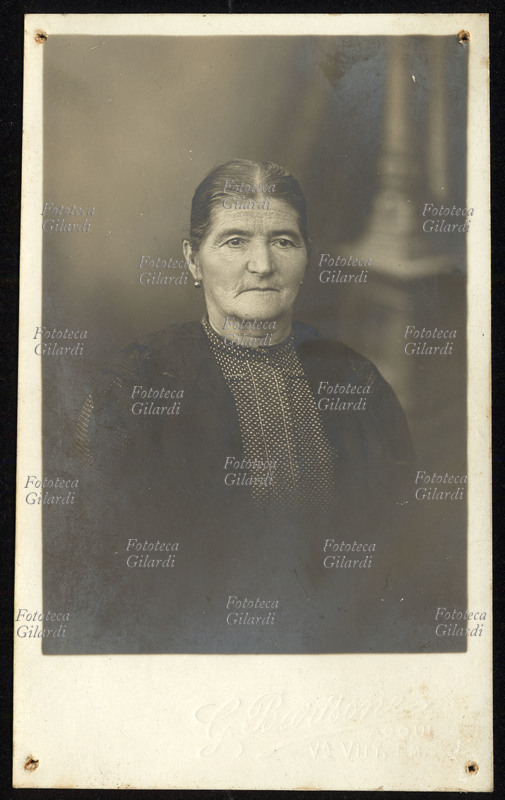 DONNA anziana ritratta a mezzo busto, vestita di giacchetta nera con camicia senza collo scura a pallini bianchi. Fotografia di anonimo, 1930 circa.