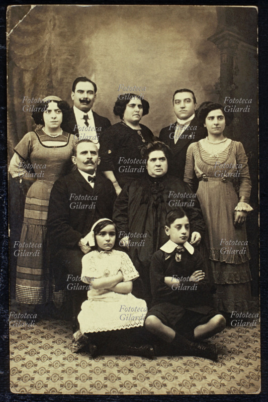 FAMIGLIA estesa, fotogruppo composto dai genitori, seduti al centro, con i figli ed i relativi coniugi. Fotografia di anonimo, eseguita in atelier. Italia, 1900 circa.