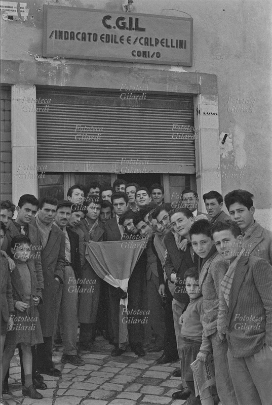 COMISO C.G.I.L. Sindacato Edile e Scalpellini. Operai davanti alla sede, la saracinesca abbassata a metà e il tricolore listato a lutto per la morte di Paolo Vitale, ucciso dalla Celere durante la manifestazione del 20 febbraio. Fotografia di Ando Gilardi (1921-2012) #andogilardi, Comiso (Ragusa), 1956.