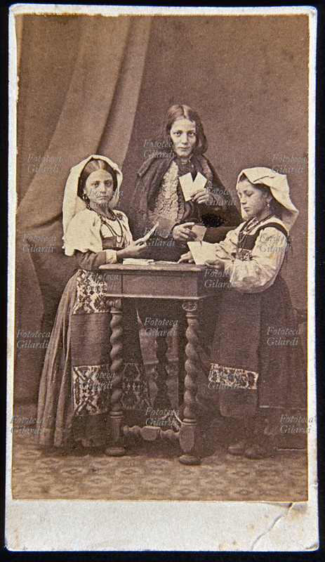 TRE BAMBINI con vestiti tradizionali campani o calabresi giocano a carte. Le due bambine sono ai lati del tavolino mentre il bambino è di fronte al fotografo. Italia, fotografia di anonimo 1880 circa.