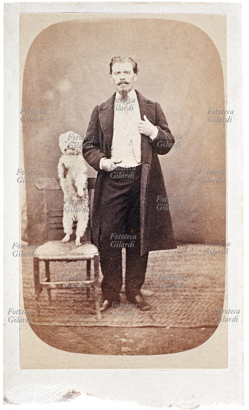 FRIENDS male full-length portrait; the man is accompanied by his faithful dog, a poodle posing "standing" on its hind legs next to him, on a chair. The picture is not very engraved because in order to capture the little dog with a fast time, the photographer had to use, probably, a very wide aperture. Photography sepia, Italy around 1900.