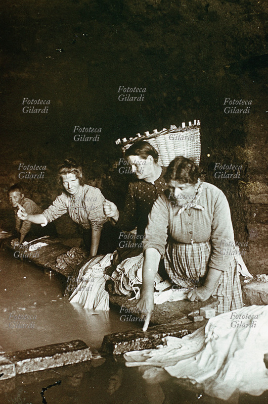 MESTIERI Lavandaie al lavoro in una vasca pubblica: i panni venivano trasportati nella gerla, come si vede, e lavati sulla pietra. Due di loro mimano il caratteristico gesto con l\