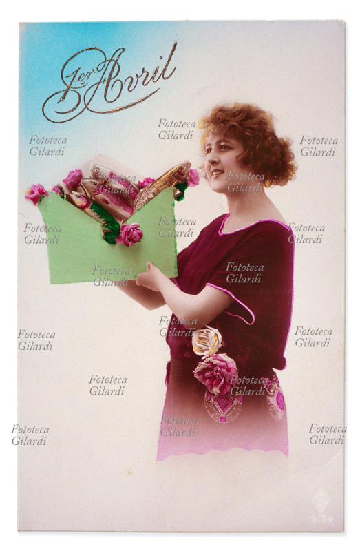 RICORRENZE Primo Aprile, giovane donna sorridente riceve una grande busta verde colma di pesci. Cartolina fotografica acquarellata. Francia, 1923