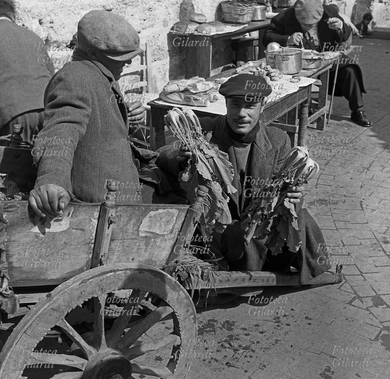 AGRICOLTORI in posa accanto al loro carretto, tipico siciliano, con in mano cime di rapa che propongono alla vendita. Sullo sfondo un banchetto che vende pane e cibo da strada in pentola, con un avventore seduto a consumare la sua porzione. Fotografia di Ando Gilardi (1921 - 2012) #andogilardi, Palermo 1955 circa.