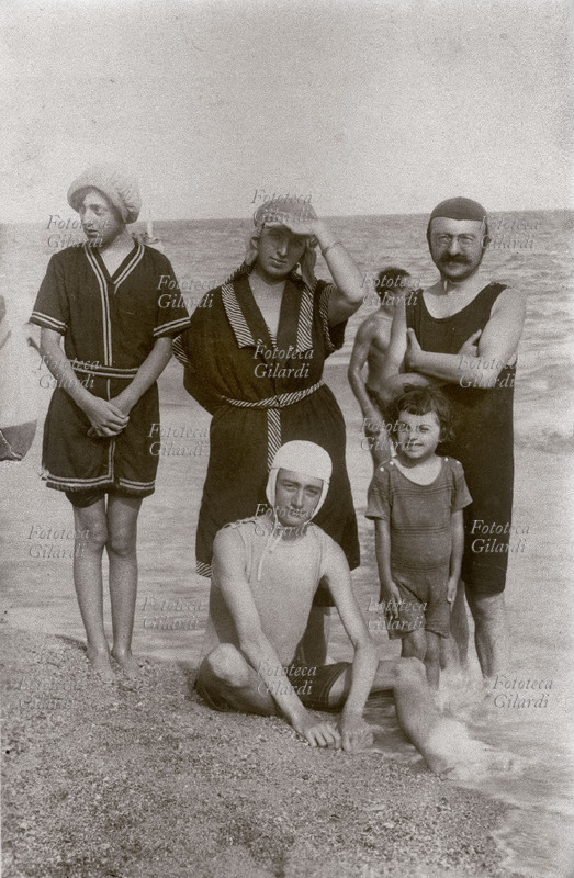 VACANZE Gruppo familiare in riva al mare, posa per una foto-ricordo. I genitori posano in piedi con le figlie femmine, mentre il figlio maschio sta seduto sul bagnasciuga. Italia, 1914-1921