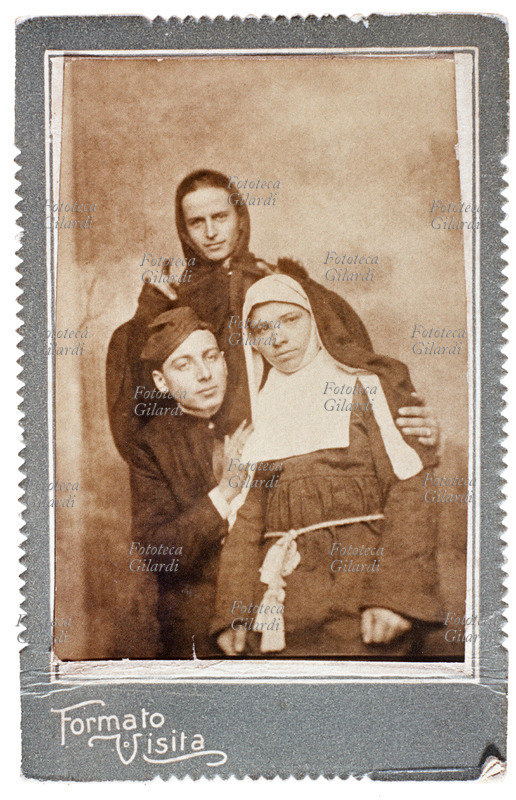TRAVESTIMENTO scherzoso di tre amici che si sono fatti fotografare in abiti da prete, suora e monaco. La "suora" siede sulle ginocchia del "prete", il "monaco" in piedi alle loro spalle sembra imporre la sua benedizione. Fotografia formato Carte de Visite, Italia 1910 circa.