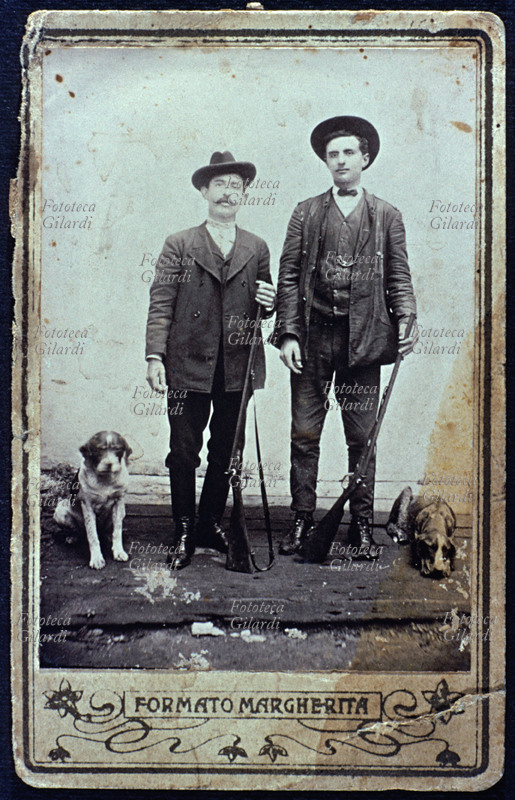 CACCIA due cacciatori in posa, a figura intera, ciascuno con fucile e cane; anche i due animali sono disciplinatamante immobili in posa. Fotografia eseguita in atelier, formato Margherita, Italia 1900 circa.