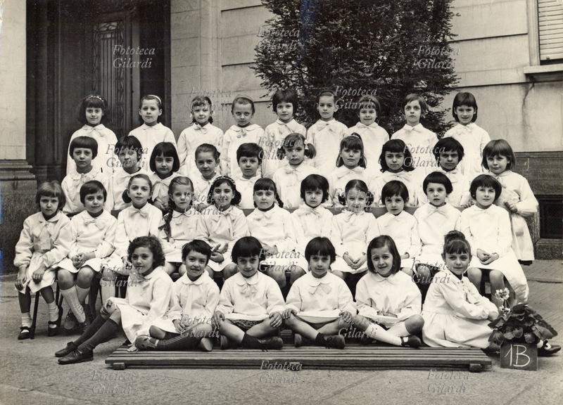 SCUOLA Fotografia ricordo di una classe femminile delle elementari: 1B. Le alunne indossano tutte il classico grembiule bianco contraddistinto da un fiocco chiaro. Al centro in prima fila una coppia di gemelle. La maestra unica, non presente nella foto, gestiva da sola l\