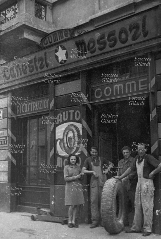 GOMMISTA negozio-officina ricostruzione gomme, in un quartiere periferico milanese. Il proprietario ed il personale in posa con un grande pneumatico da camion. Milano, 1945.