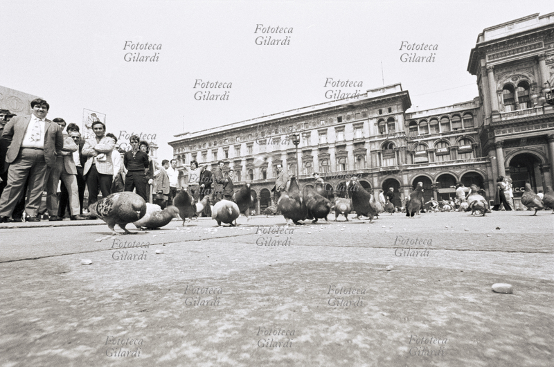STORIA DELLA FOTOGRAFIA veduta di piazza del Duomo a Milano con i piccioni che beccano il mangime lanciato dai turisti. Insolita veduta rasoterra, presa con un apparecchio Hologon su treppiede con autoscatto; un capannello di curiosi si è fermato ad osservare il fotografo all\