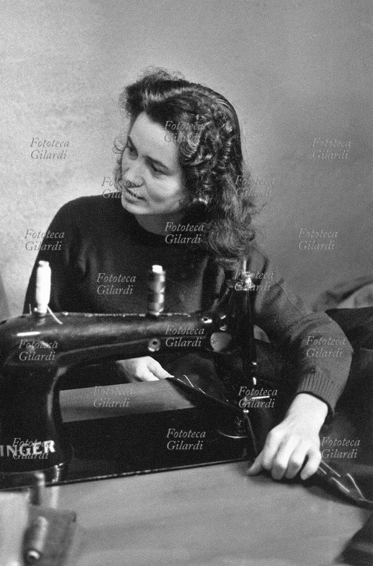 MESTIERI Allegra sarta al lavoro alla macchina da cucire; la giovane sorride e si gira graziosamente verso la collega, distogliendo un momento lo sguardo dal lavoro; cuce con una delle prime \