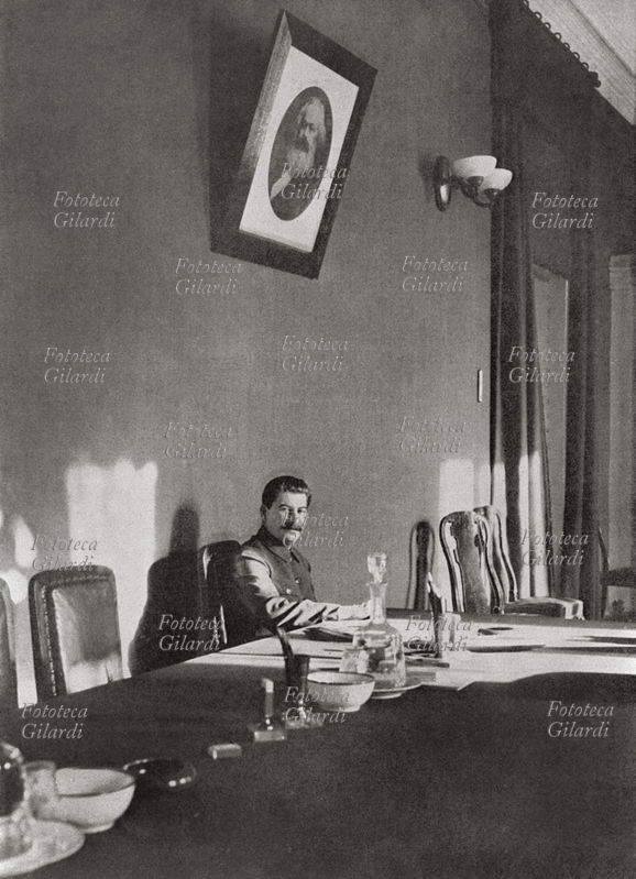 STALIN (1879-1953) seduto al tavolo delle riunioni, sotto un ritratto di Karl Marx incombente nell\