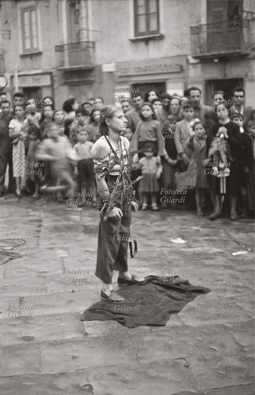 ILLUSIONISMO giovane donna, artista girovaga di piazza, si faceva legare stretta da un assistente con catene e spade, per poi slegarsi alla maniera del celebre mago Houdini sotto gli occhi del pubblico, appena se ne era radunato a sufficienza fra i passanti. Fotografia di Ando Gilardi (1921 - 2012) #andogilardi, Crotone 1957.Questa immagine è inclusa nel nostro fotolibro in vendita qui.