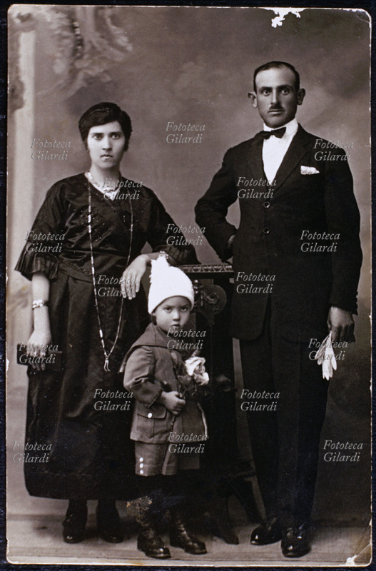 FAMIGLIA genitori in posa con il figlioletto. Entrambi sono elegantemente vestiti, la madre porta numerosi gioielli, ma l\