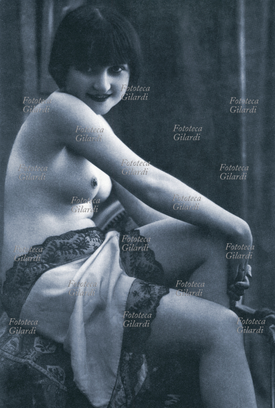 SEDUTTRICE Donna a seno nudo sorride teneramente, seduta si abbraccia un ginocchio. Cartolina postale, circa 1920