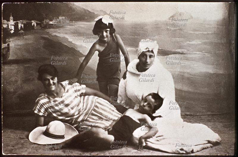 VACANZE AL MARE Fotografia ricordo di bagnanti realizzata nello studio di un fotografo. Due mamme con i due figlioletti posano negli abiti da spiaggia: le signore con accappatoio e prendisole; i bambini in costume da bagno. Italia, 1920 - 1925