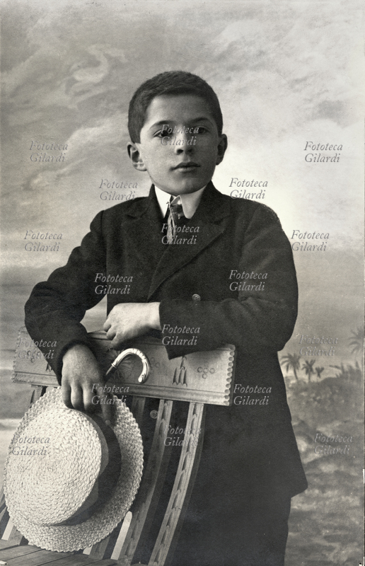 BAMBINI Bambino di circa 10 anni posa per una fotografia ricordo appoggiato alla spalliera di una sedia, in atteggiamento da piccolo gentiluomo; in mano tiene il cappello (paglietta) e il bastone da passeggio. Italia, primi del Novecento