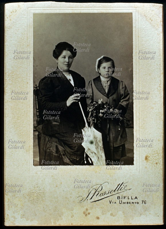 FAMIGLIA madre e figlia di età intorno ai sette anni. La donna è seduta e la bambima in piedi, tutte e due eleganti, in abito da cerimonia, si assomigliano nei tratti somatici. Fotografia di S. Rossetti, eseguita in atelier. Biella, 1915 circa.