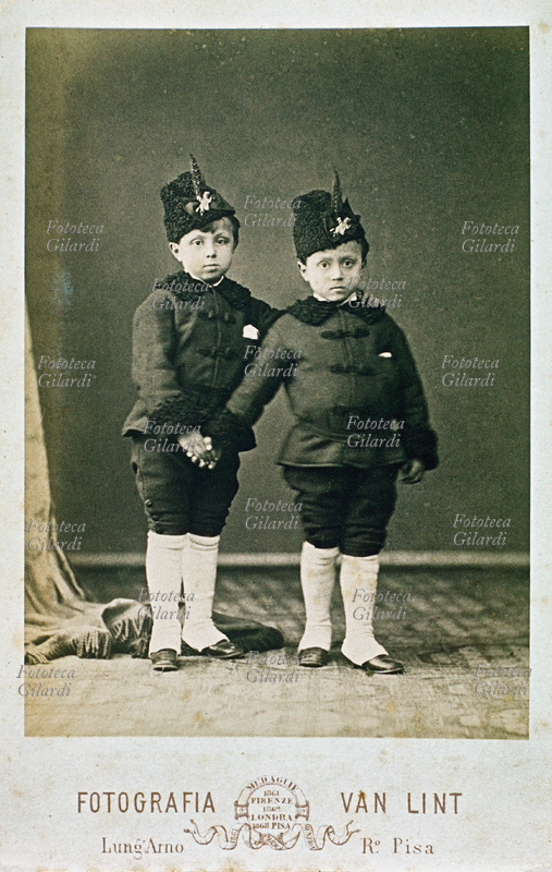 BAMBINI Fotografia ricordo. Due fratellini (5-6 anni) posano per mano, in divisa con ghette bianche e colbacco con piuma. Fotografia Van Lint, Pisa, 1880 circa.