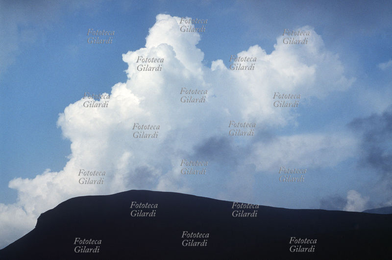 NUVOLA bianca spumosa sale da dietro la sagoma di una montagna, nel cielo azzurro. Un cumulo: nube isolata bianca e densa con sommità a forma di cupola. Si origina per risalita di masse d\