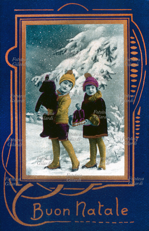 NATALE auguri. Due bambini in un paesaggio innevato, sotto un abete carico di neve, porgono gli auguri con un sorriso, stringendo un orso di pezza e una bambola meccanica, appena ricevuti in dono. Cartolina postale fotolitografica monocromatica colorata a pouchoir e dorata, Italia circa 1930.