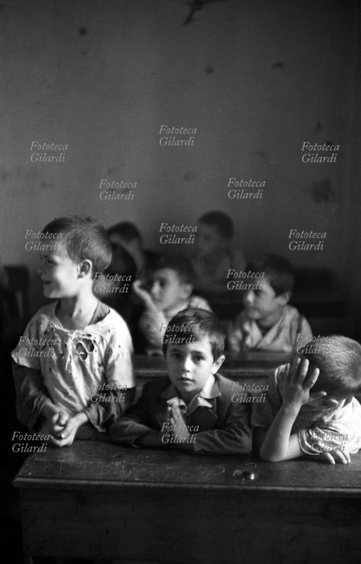 SCUOLA aula in classe mista, i bambini in età da scuola elementare (intorno ai sette anni) seguono la lezione di fronte alla maestra. Sono seduti nei banchi a tre a tre senza badare alle differenze; nel settore maschile stanno fianco a fianco un bimbo che indossa la giacchetta \