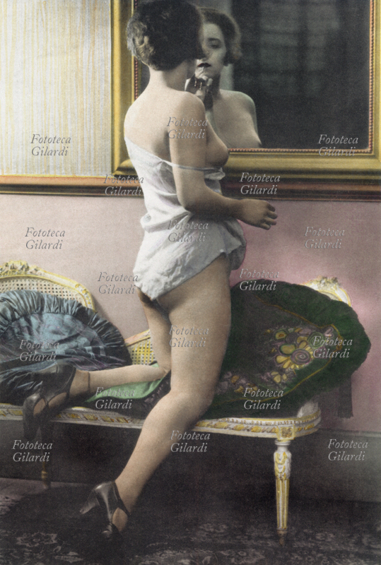 EROS giovane donna in sottoveste davanti allo specchio, si ritocca il trucco; fa pensare che si prepari a esercitare la sua professione di seduttrice, in una casa chiusa. Fotografia, primi anni del XX secolo