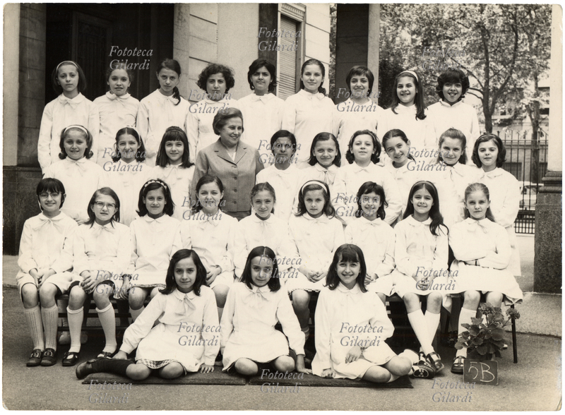 SCUOLA Fotografia ricordo di una classe femminile delle elementari: 5B. Le alunne indossano tutte il classico grembiule bianco contraddistinto da un fiocco chiaro. In prima fila al centro due gemelle. Le bambine sorridono tutte o quasi, così come la maestra che sorride ma che al contrario delle sue allieve, non guarda in macchina. Il clima è allegro, addirittura una bambina monella tira fuori la lingua in espressione beffarda. La maestra unica gestiva da sola l\