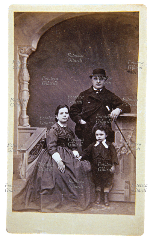 FAMIGLIA in posa sotto un arco, nello studio di un fotografo. La giovane madre è seduta e tiene per mano il figlioletto. Il padre è in piedi appoggiato al finto parapetto e indossa una bombetta, in mano tiene un bastone da passeggio. Italia, 1885 ca.
