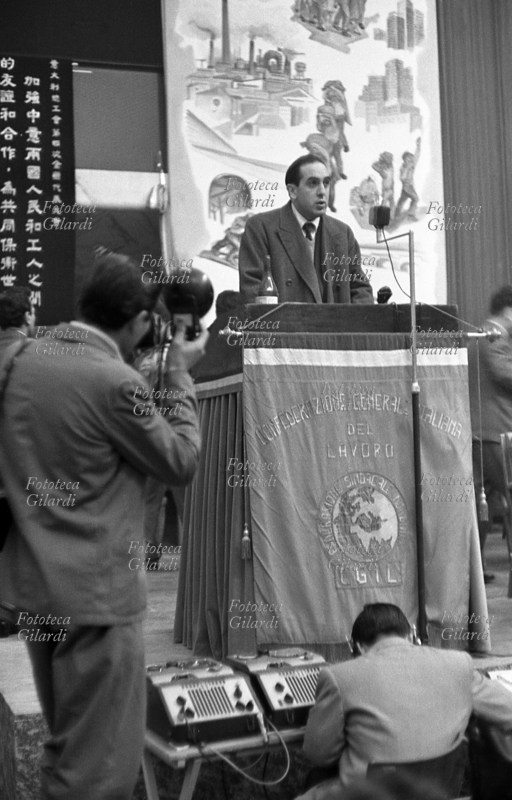 IV CONGRESSO della CGIL riunito a Roma EUR. Un relatore sul palco parla alla sala gremita di pubblico, un fotografo lo riprende e un fonico registra. Sullo sfondo un drappo con scritte in cinese. Fotografia di Ando Gilardi (1921 - 2012), #andogilardi, Roma, 27 febbraio/4 marzo 1956.