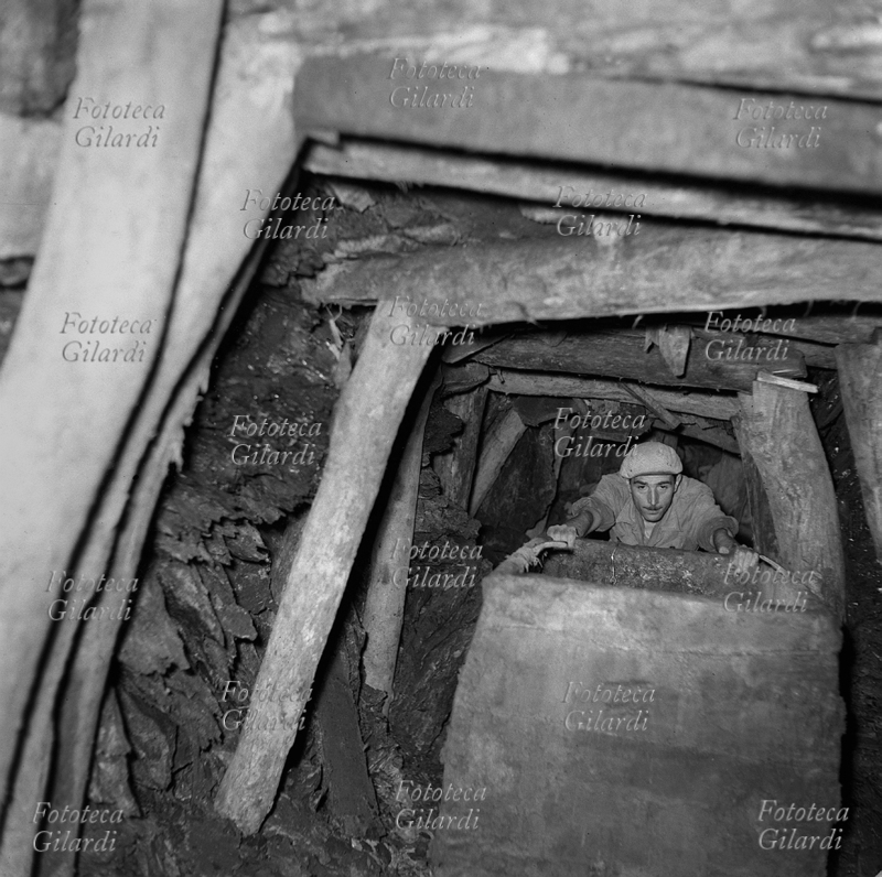MINATORE spinge a mano un carrello nella antichissima miniera di lignite a Luni, detta la Lunara, da cui si estraeva già al tempo degli Etruschi. La lignite è un carbone fossile con caratteristiche di combustibile poco pregiate, entrato in disuso nel secondo dopoguerra (1950-60). Gli operai occuparono questa miniera nel tentativo di impedirne la chiusura, che poi invece avvenne con conseguente licenziamento dei lavoratori. Fotografia di Ando Gilardi (1921-2012) #andogilardi, eseguita con illuminazione di bulbi al magnesio. Luni (La Spezia), 1953.