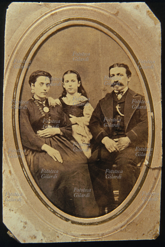 FAMIGLIA genitori e figlia di età intorno ai dieci anni. I due adulti sono seduti e la mambina è dietro di loro in piedi. Il ritratto è montato in un passe partout ovale. Fotografia di anonimo eseguita in atelier, Italia 1880 circa.
