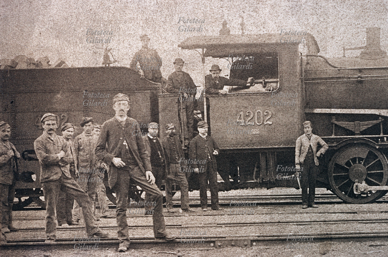 TRENI personale addetto alla manovra ed alla manutenzione ferroviaria in posa sui binari, vicino ad un convoglio con locomotiva (v. 43141 per dettaglio oliatore). Italia, 1880 circa.
