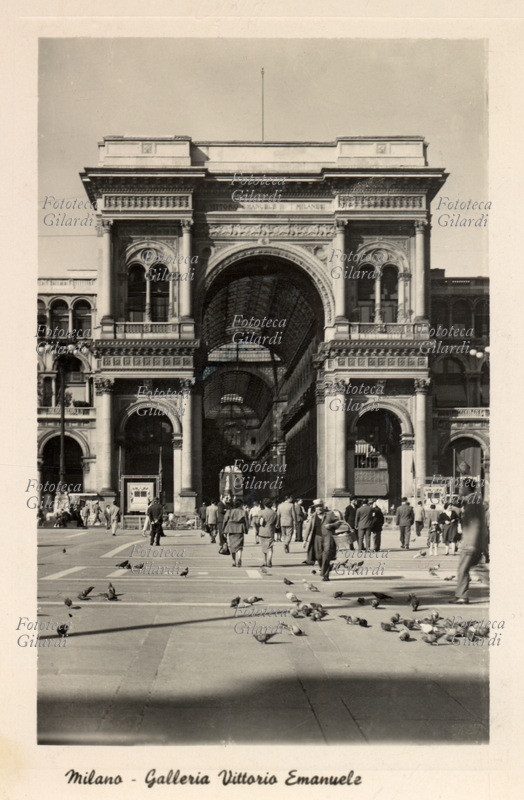 MILANO Galleria Vittorio Emanuele. Ingresso della galleria da piazza del Duomo, in primo piano alcuni piccioni che popolano la piazza becchettano indisturbati sul selciato; al centro un venditore ambulante di mangime per uccelli con cesto. Cartolina postale, fotografia, Milano 1955.