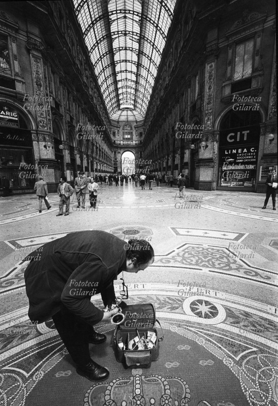 Ando GILARDI (1921-2012) ritratto a Milano in Galleria Vittorio Emanuele, mentre si appresta ad estrarre la sua apparecchiatura fotografica per un servizio. Il ritratto è eseguito con grandangolare. Fotografia di Luciana Barbarino Gilardi, 1969 circa