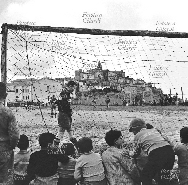 CALCIO campo di calcio alle porte di Albano di Lucania (Potenza), partita tra squadre giovanili. Ripresa a bordo campo da dietro la porta, da dove i bambini osservano e commentano le azioni accovacciati per terra; sullo sfondo, il paese. Fotografia di Ando Gilardi (1921 - 2012) #andogilardi, Italia 1957.