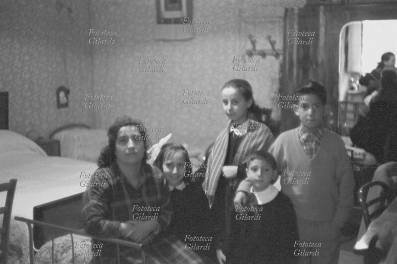 FAMIGLIA calabrese. In camera da letto una madre in posa con i suoi quattro figli (due maschi e due femmine). I tre più piccoli indossano il grembiule della scuola. Sullo specchio dell\