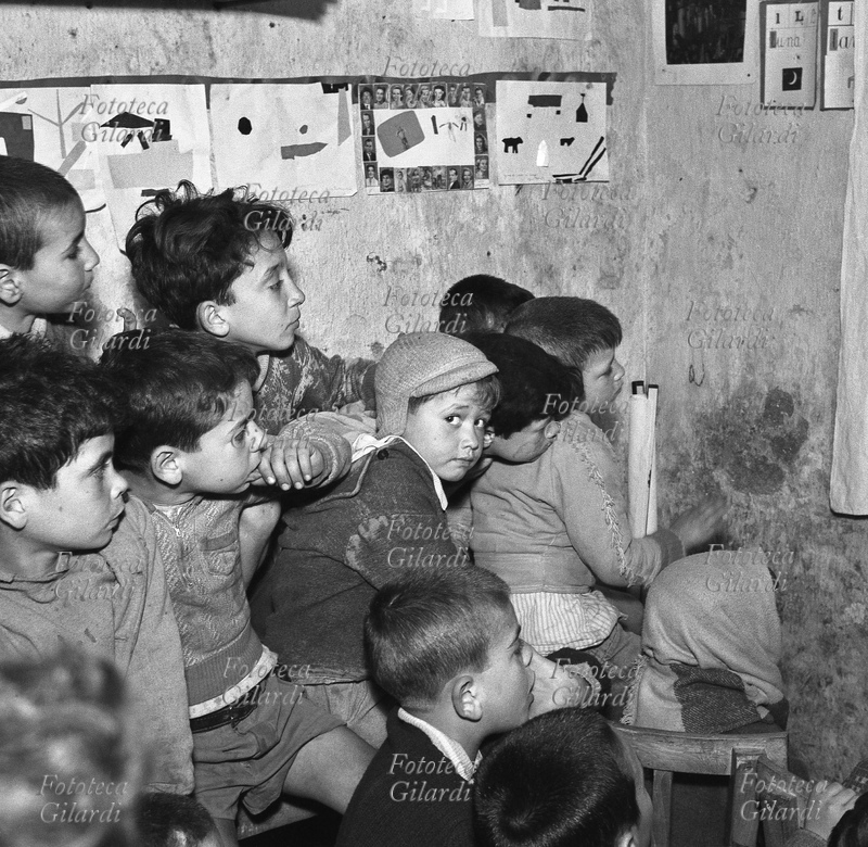 DOPOSCUOLA a cortile Scalilla, tutti gli scolari attenti alla lezione, tenuta in questo caso sottoforma di piccolo spettacolo di pupazzi, tranne un bambino al centro che guarda in macchina; lezione tenuta dai coniugi Michela e Goffredo Fofi. Fotografia di Ando Gilardi (1921 - 2012) #andogilardi da ampio servizio [vedi anche 17117]. Palermo, 1957.