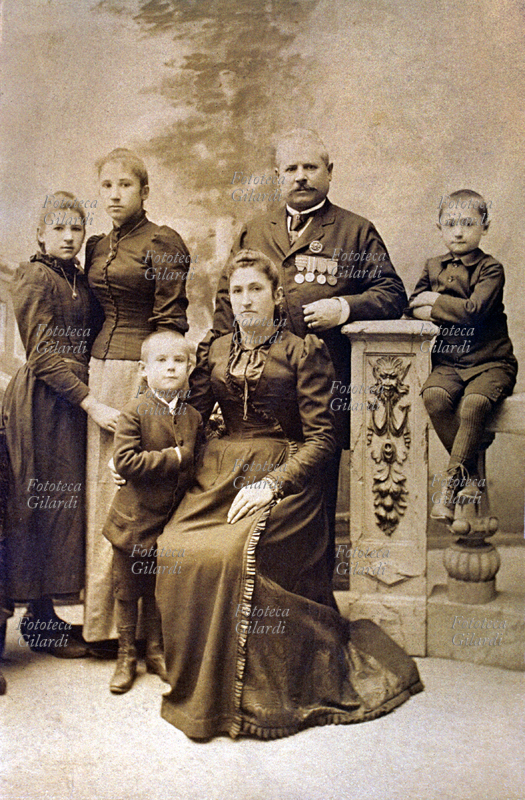 FAMIGLIA genitori con quattro figli posano nello studio del fotografo per una fotoricordo. Seduta al centro del gruppo la madre con abito lungo e scuro. Dietro a lei (in piedi) il marito pluridecorato con medaglie appuntate sul petto. I figli sono due maschi (più giovani) e due femmine. Italia, 1890 circa