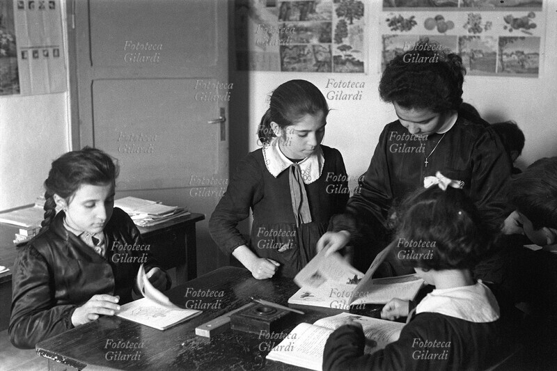 SCUOLA Lezione in una classe elementare: la maestra sceglie la lettura mentre le scolare cercano la pagina sul loro libro. Fotografia di Ando Gilardi (1921 - 2012) #andogilardi, Polino (Terni), 1958 circa.