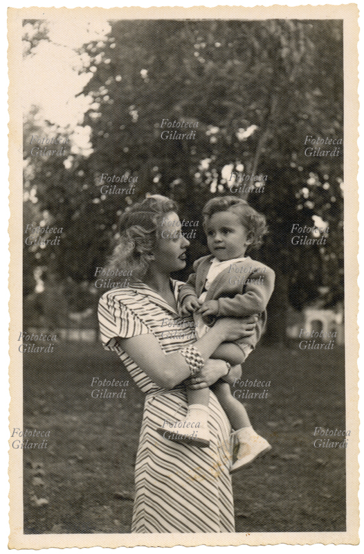 MAMMA con bambina in braccio, di età intorno ai due anni, in un parco giardino. Fotografia su carta millepunti, Novi Ligure (AL) 1950
