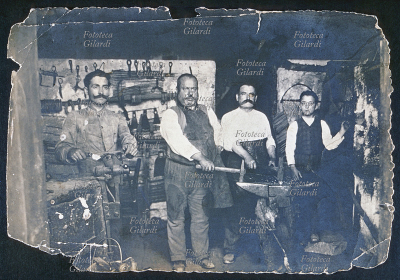 MESTIERI FABBRO FERRAIO posa per una foto ricordo insieme al figli e un giovane apprendista. Alcuni prodotti sono apppesi sulla parete di fondo, zappe e grosse forbici. Indossano diversi modelli di grembiule. Rara fotografia di un artigiano di altri tempi, 1902 circa
