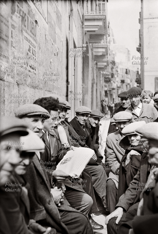REPRESSIONE in una strada di Barletta un giovane legge ad alta voce sul giornale la notizia dei fatti del 14 marzo 1956: le forze di polizia aprirono il fuoco contro circa 4.000 persone esasperate dalla fame che manifestavano contro le discriminazioni della Pontificia Opera di Assistenza, incaricata dall\