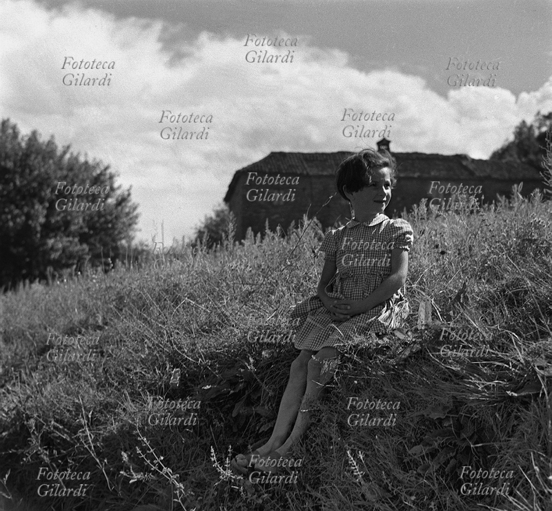 BAMBINA ritratta sul bordo di un campo, sullo sfondo di una cascina rurale tra le colline del basso Piemonte, in provincia di Alessandria. Fotografia di Ando Gilardi (1921 - 2012) #andogilardi, Italia 1952.